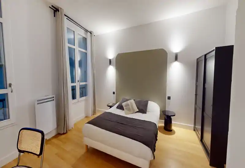 Chambre moderne et lumineuse avec lit double centré, tête de lit stylée et tables de chevet ; parquet chaleureux et grandes fenêtres apportant de la lumière naturelle.