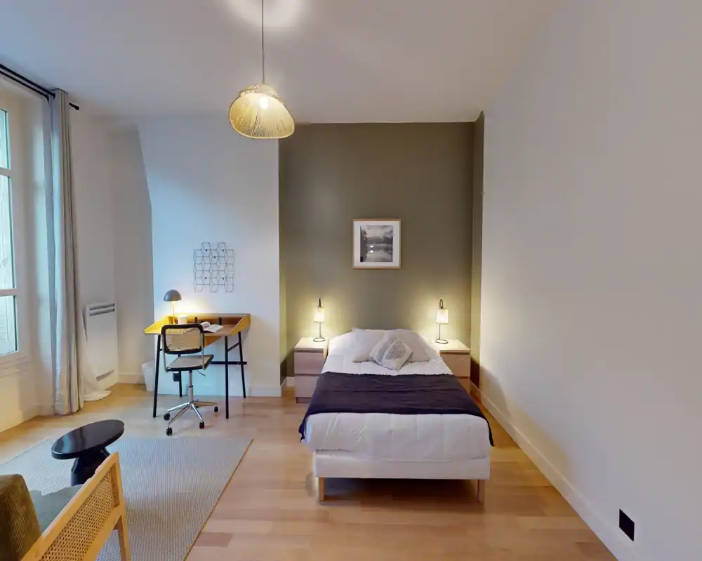 Chambre lumineuse et spacieuse avec un lit simple centré sur un mur vert, tables de chevet avec lampes, un petit bureau près de la fenêtre et un sol en bois chaleureux.