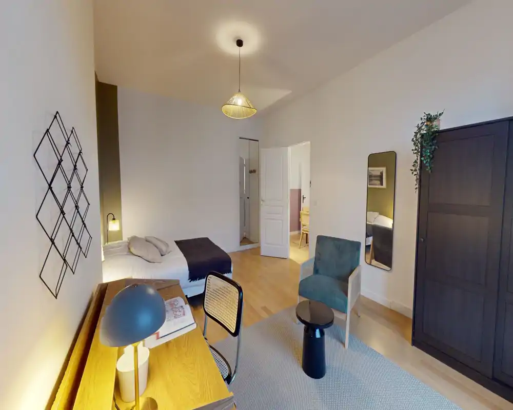 Chambre lumineuse et élégante avec lit double, bureau et coin salon ; lumière chaleureuse et armoire pour un séjour confortable et bien aménagé.