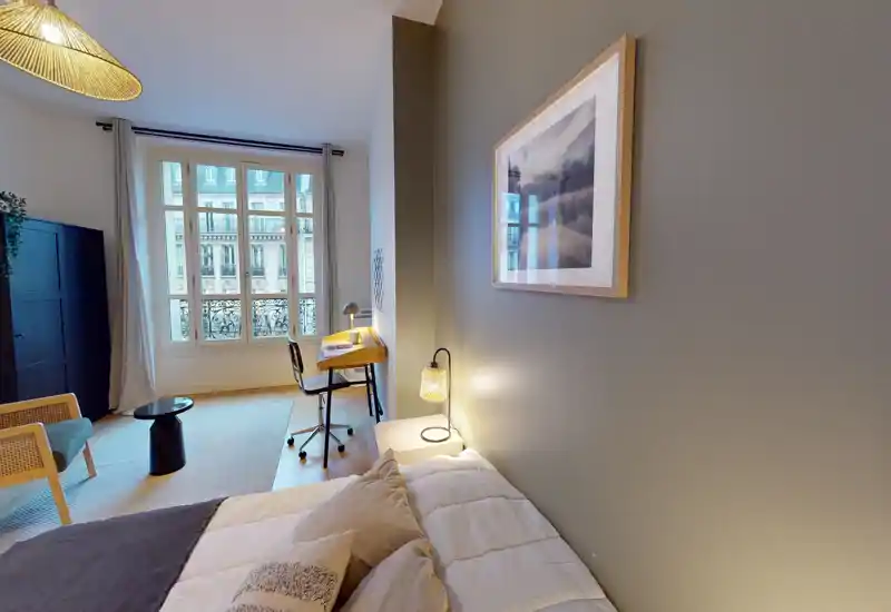 Chambre lumineuse et élégante avec un lit confortable au premier plan, un bureau près d'une grande fenêtre et une lampe de chevet chaleureuse — idéale pour travailler et se détendre.