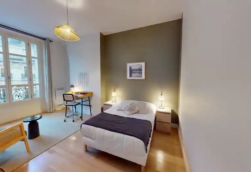 Chambre lumineuse et bien décorée avec un lit double encadré par des tables de chevet et lampes, un bureau près de la fenêtre et un sol en bois chaleureux — idéale pour les annonces de location.