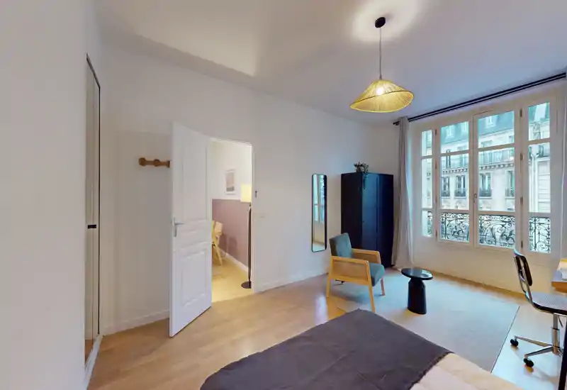 Chambre lumineuse et agréablement meublée avec un lit double au premier plan, un fauteuil, un bureau près d'une grande fenêtre et un éclairage soigné — idéale pour un séjour en ville.