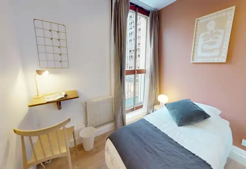 Compact, bright bedroom with a neatly made single bed, bedside lamp and a small workspace by the window. Warm accent wall and natural light make the room welcoming for short stays.