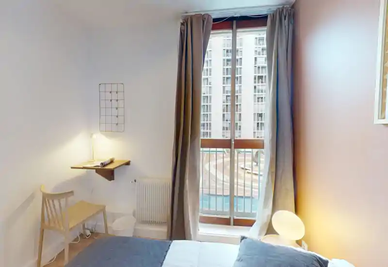 Compact, bright bedroom with a double bed, bedside lamp and a large window with curtains overlooking the courtyard. Minimal wooden chair and small wall shelf create a functional, cozy corner ideal for short stays.