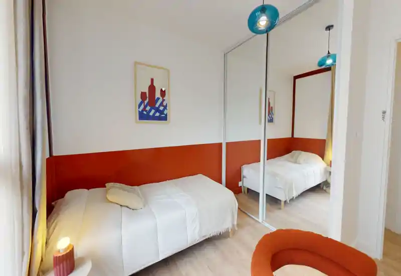 Bright, neatly styled single bedroom with a white bed, warm orange accent wall and mirrored wardrobe that adds depth. Soft linens, bedside lighting and a cosy chair make it ideal for a comfortable stay.