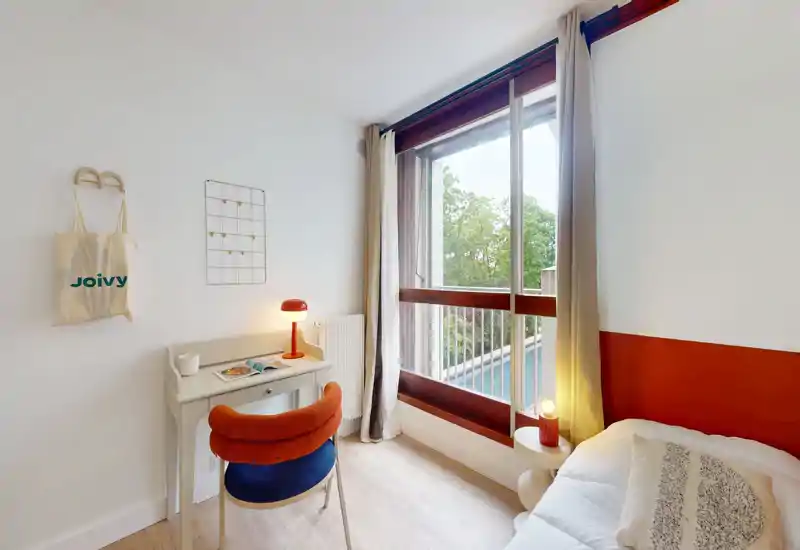 A bright, modern bedroom corner with a comfortable single bed, bedside table, and a small desk by a large window offering leafy views — cozy, well-lit and styled for rental photos.