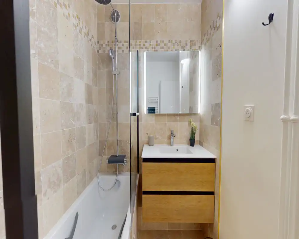 Modern compact bathroom with a bathtub and overhead shower, wood-finish vanity with integrated sink, illuminated mirror and beige stone-effect tiles.