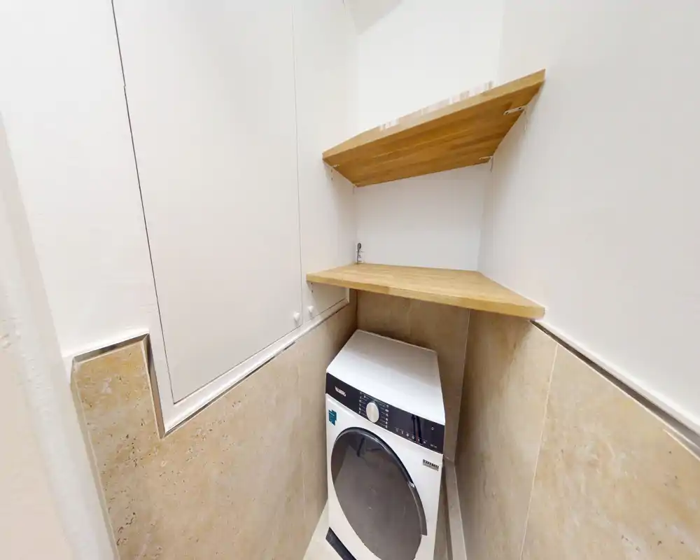 Compact laundry/utility alcove with a front-loading washer and two wooden shelves above — a neat service corner suitable for functional storage.
