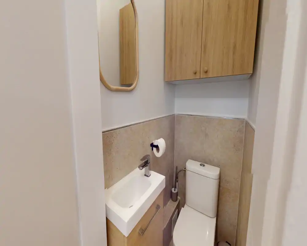 Compact, well-lit bathroom with a toilet, a small rectangular sink with faucet, wall-mounted mirror and wooden storage cabinet.