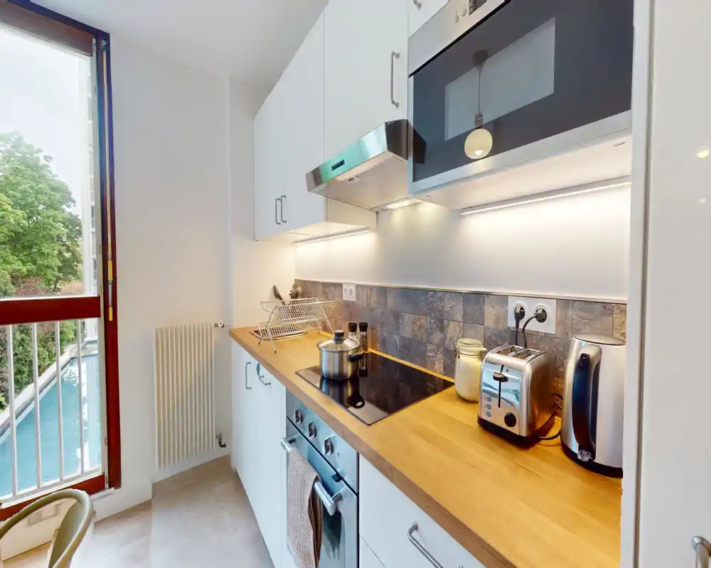 Modern compact kitchen with a wooden countertop, induction hob, built-in oven, toaster and kettle; bright natural light from a large window and integrated under-cabinet lighting.