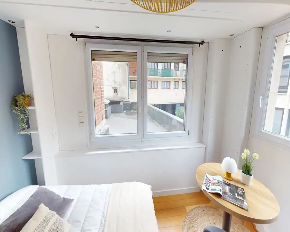 Dormitorio compacto y luminoso con cama confortable, luz natural por una ventana amplia y una pequeña mesa de madera con objetos decorativos — ideal para estancias acogedoras.