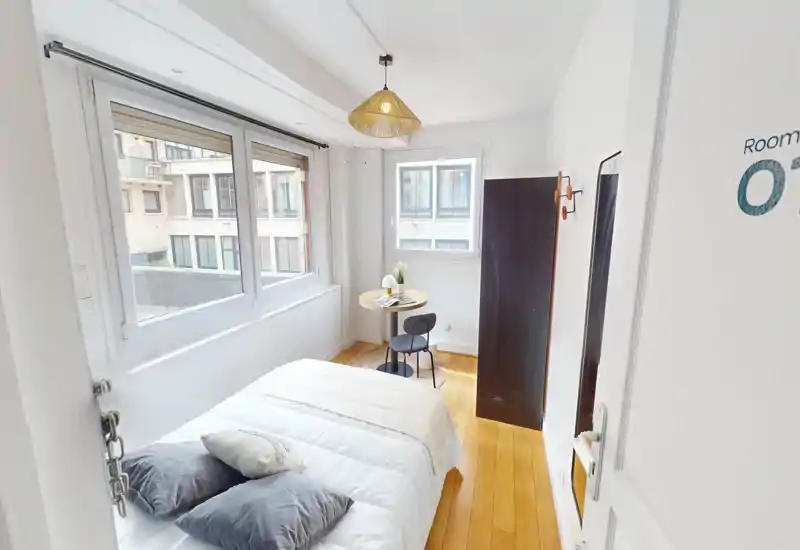 Dormitorio luminoso y acogedor con cama doble, cojines suaves y luz natural de grandes ventanas; pequeña mesa redonda y silla junto a la ventana para un espacio de trabajo compacto.
