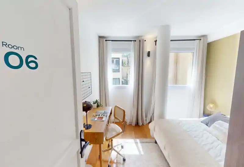 Camera da letto luminosa e ben arredata con un comodo letto matrimoniale, luce naturale da due finestre e una piccola scrivania in legno — perfetta per riposo o lavoro da remoto.