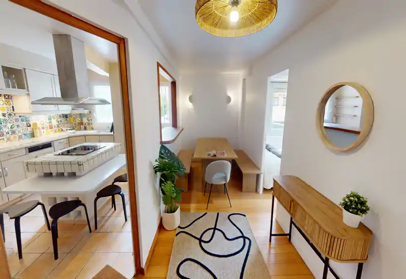 Coin salon/salle à manger lumineux et étroit avec petite table en bois, banquettes et vue sur une cuisine carrelée. Parquet chaleureux, tapis décoratif et plantes pour une ambiance accueillante.