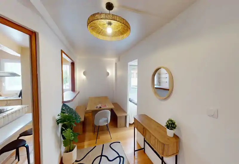 Coin repas chaleureux et étroit avec table en bois, banc et suspension décorative ; la lumière naturelle venant de la cuisine crée une atmosphère accueillante.
