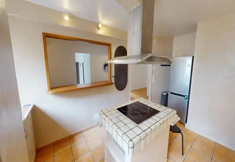 Cuisine compacte équipée avec îlot central carrelé, plaque vitrocéramique et hotte suspendue ; carrelage chaleureux et passe-plat vers la pièce adjacente.