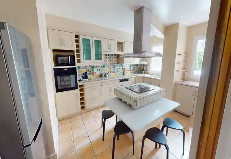 Cuisine lumineuse et bien équipée avec crédence de carreaux colorés, four et réfrigérateur encastrés, îlot central avec plaque de cuisson et sièges décontractés — parfaite pour cuisiner en famille et recevoir.
