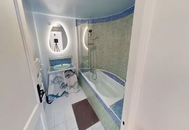Salle de bains lumineuse carrelée avec baignoire et douche, petit lavabo avec rideau décoratif, miroir éclairé et murs en mosaïque.