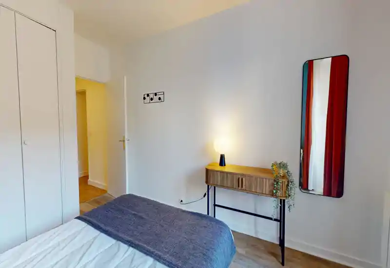 Chambre lumineuse au style épuré avec un lit bien fait, une console en bois avec lampe et plante, et un miroir en pied. Lignes nettes et textiles chaleureux créent une atmosphère accueillante pour une annonce.