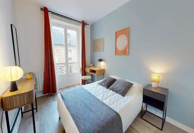 Chambre lumineuse et stylée avec lit double, tables de chevet et éclairage chaleureux. Mur d'accent bleu pâle, sol en bois et grande fenêtre avec rideaux couleur rouille laissant entrer la lumière naturelle.