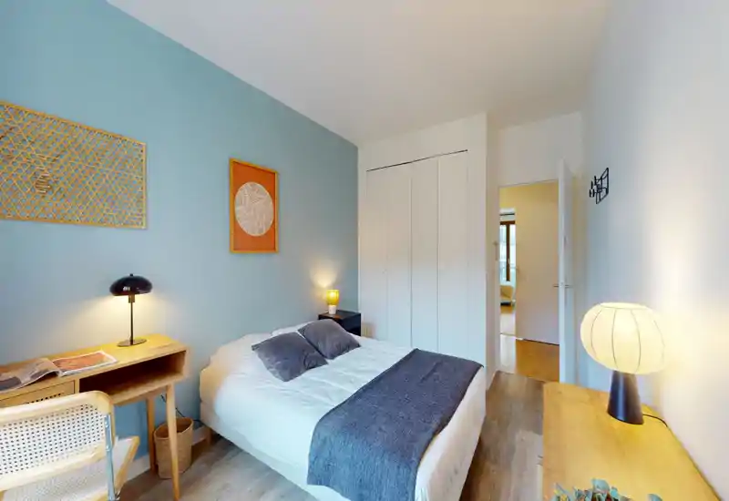 Chambre lumineuse et moderne avec lit double, tables de chevet et lampes chaleureuses, petit bureau et placard intégré — idéale pour un séjour confortable.