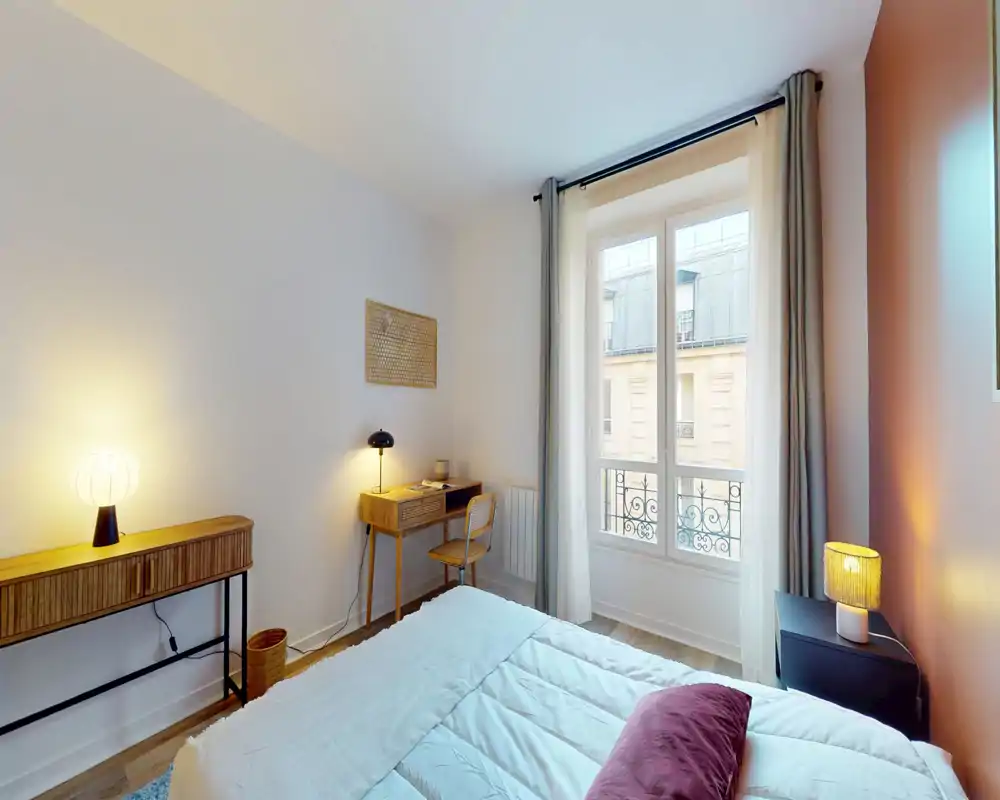 Chambre lumineuse et compacte avec lit double, linge de lit doux, table de chevet et bureau près de la fenêtre. La lumière naturelle entre par les grandes fenêtres et des lampes chaudes ajoutent une ambiance cosy.