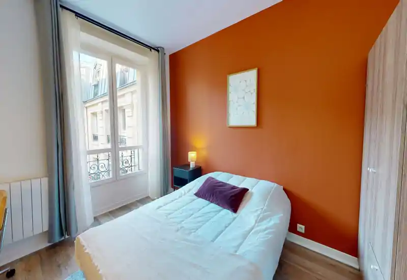 Chambre lumineuse et chaleureuse avec lit double, mur d'accent orange et grande fenêtre apportant une belle lumière naturelle. Idéale pour les locations de courte durée cherchant un espace nuit stylé et confortable.
