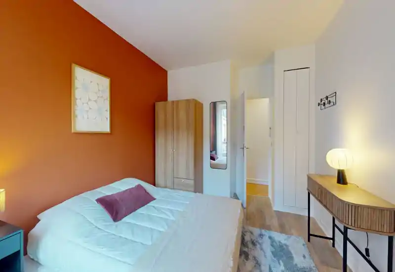 Chambre lumineuse et moderne avec un mur d'accent terre cuite, lit double avec literie blanche et coussin décoratif, armoire, miroir en pied et petit bureau avec lampe.