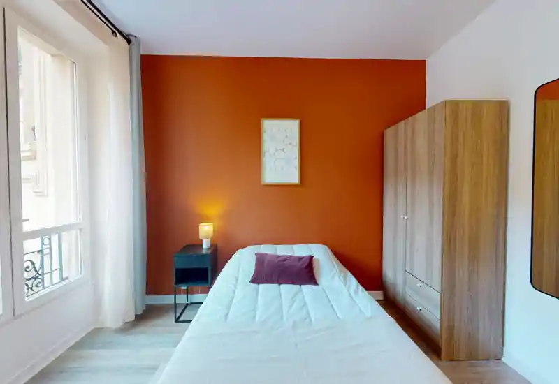 Chambre lumineuse et cosy avec lit simple central, mur d'accent terracotta, table de chevet avec lampe, armoire et grande fenêtre apportant la lumière naturelle.