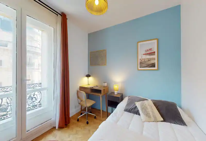 Chambre lumineuse et élégante avec un lit confortable, des textiles doux et un petit bureau en bois près de la fenêtre ; de grandes fenêtres françaises apportent une belle lumière naturelle et des lampes chaudes créent une atmosphère cosy.