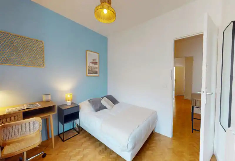 Chambre lumineuse et bien décorée avec lit double devant un mur bleu, table de chevet avec lampe et petit bureau en bois — idéale pour les annonces locatives mettant en avant le confort et un espace de travail.