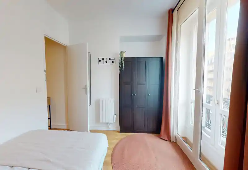 Chambre lumineuse et sobre avec lit au premier plan, une armoire foncée et de grandes fenêtres aux rideaux couleur rouille laissant entrer une abondante lumière naturelle.