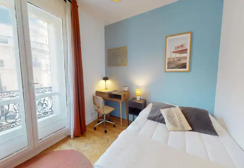 Chambre lumineuse et décorée avec un lit double confortable, une table de chevet et un petit bureau en bois près d'une grande fenêtre ouvrant sur un balcon. Textiles doux, éclairage chaleureux et un mur d'accent bleu pastel créent une ambiance cosy.