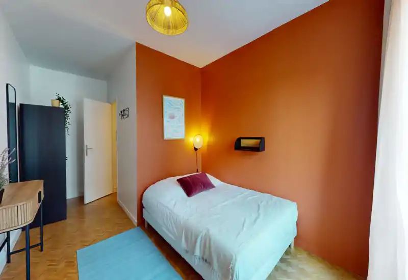 Chambre simple et chaleureuse avec un lit confortable, un mur d'accent chaleureux et une douce lumière naturelle provenant de la fenêtre. La pièce comprend une lampe de chevet, un coussin décoratif et un sol en bois soigné.