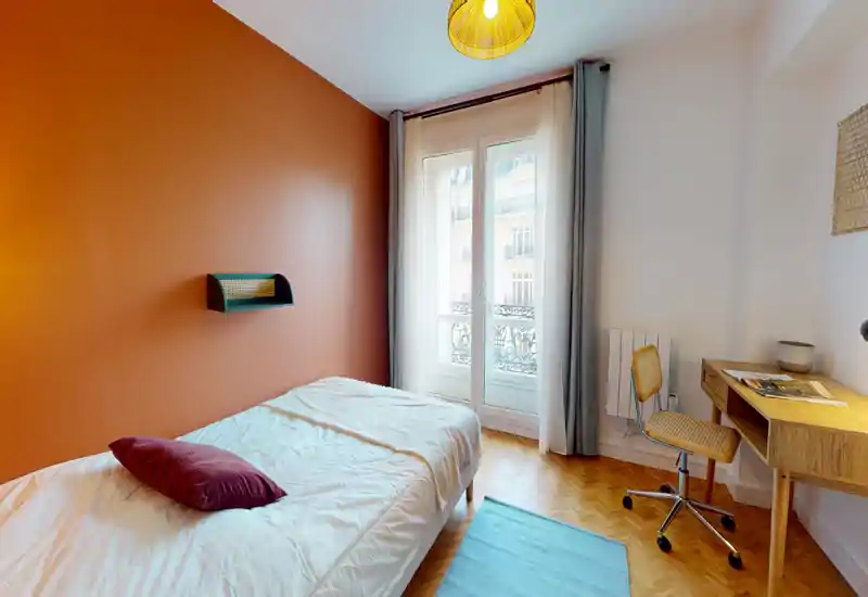 Chambre simple lumineuse et chaleureuse avec un mur couleur terre cuite, lit confortable, bureau et lumière naturelle depuis une fenêtre balcon — idéale pour étudiant ou professionnel.