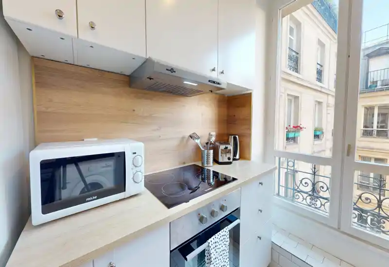 Kitchenette moderne et compacte avec plaque vitrocéramique, four, micro-ondes et crédence en bois chaleureux. Grande fenêtre apportant une belle luminosité et vue urbaine.