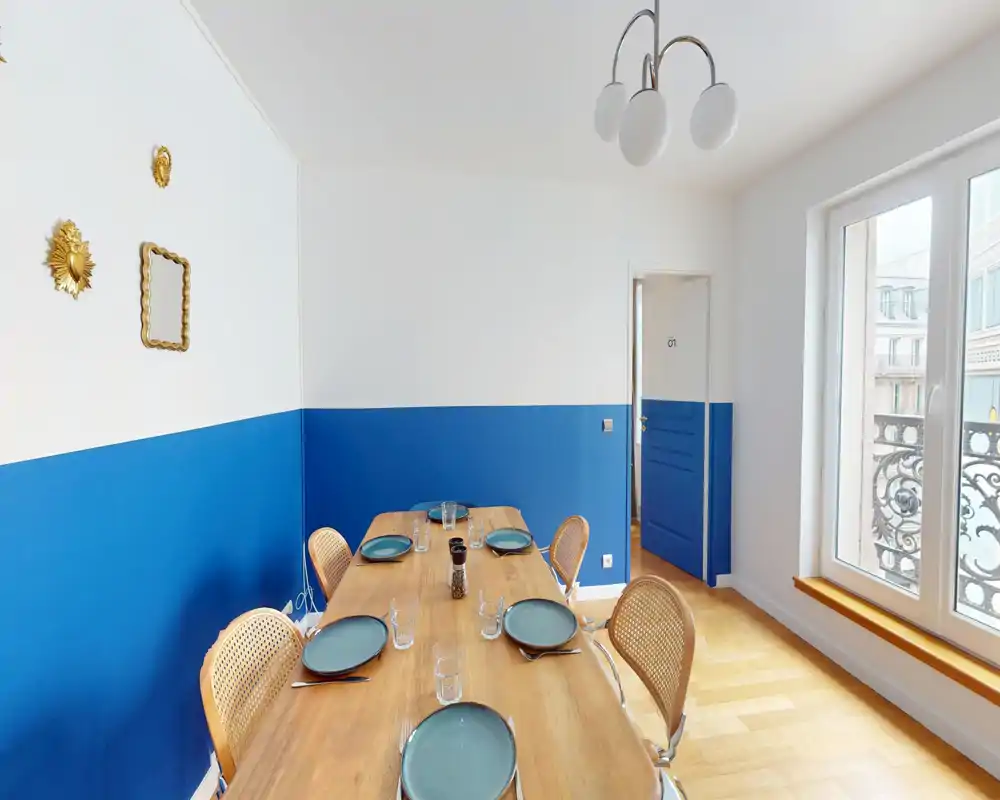 Salle à manger claire et soignée avec une grande table en bois pour six personnes, lumière naturelle par une grande fenêtre et une décoration murale bleu et blanc élégante.