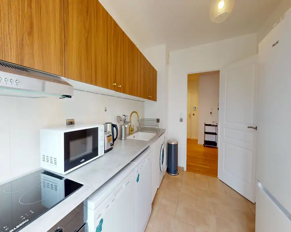 Cuisine moderne et compacte avec long plan de travail, plaque à induction, micro-ondes, évier et placards en bois suspendus. Lumineuse et bien rangée, adaptée aux locations courtes ou aux petits appartements.