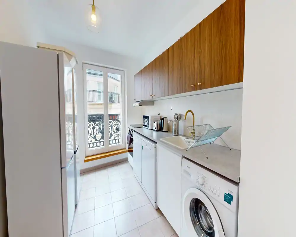 Cuisine étroite et lumineuse avec des placards hauts en bois, des appareils blancs et une grande fenêtre apportant de la lumière naturelle ; compacte mais bien équipée avec machine à laver, réfrigérateur et petits appareils sur le plan de travail.