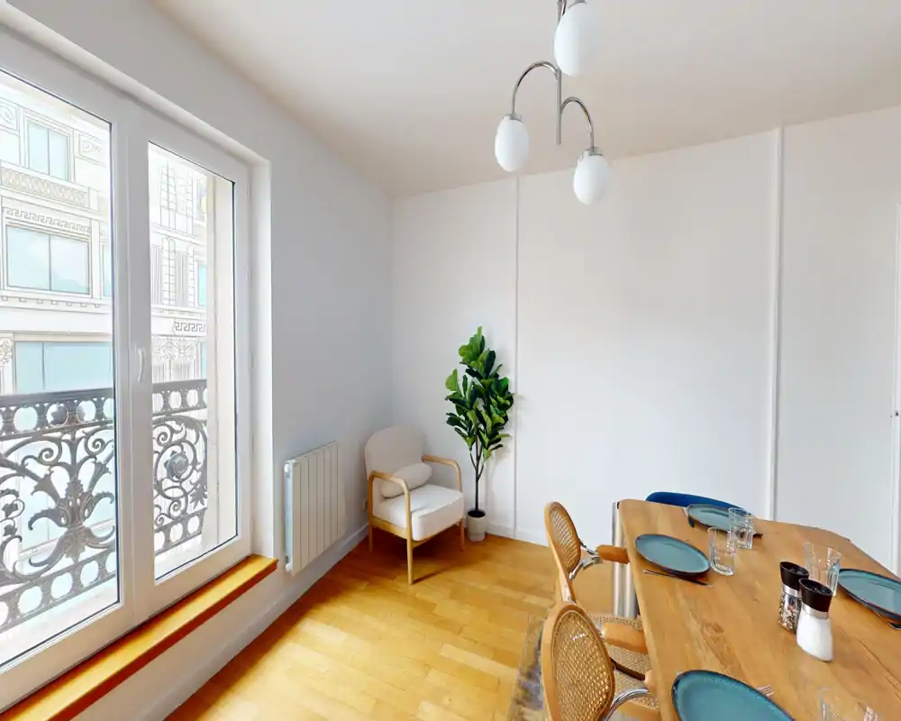 Espace repas lumineux avec une table en bois dressée pour quatre, chaises en rotin et un fauteuil près de la fenêtre ; grandes fenêtres à la française apportant une belle lumière naturelle.