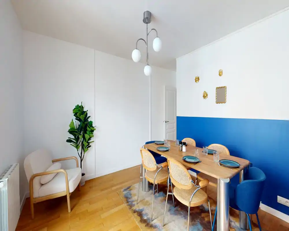 Salle à manger lumineuse et moderne avec une table en bois pour six, chaises tressées et un mur accent bleu, créant un espace élégant et accueillant.