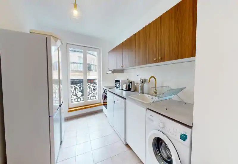 Cuisine étroite et lumineuse avec des placards hauts en bois, des appareils blancs et une grande fenêtre apportant de la lumière naturelle ; compacte mais bien équipée avec machine à laver, réfrigérateur et petits appareils sur le plan de travail.