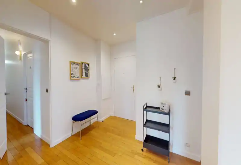 Bright and tidy entryway with wooden parquet flooring, a small blue bench and a slim black shelving trolley — welcoming, minimal decor and good natural/ambient lighting.