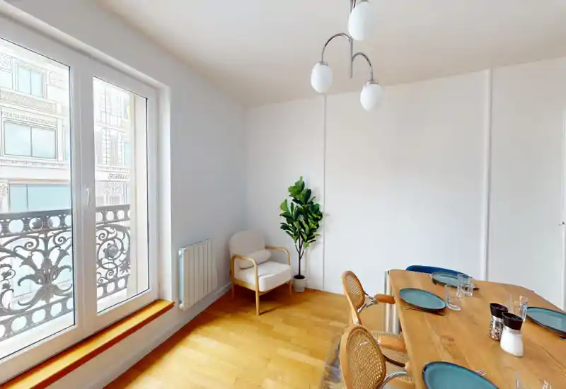 Sunlit dining area with a wooden table set for four, rattan chairs and a cozy armchair by the window; large French-style windows provide abundant natural light.