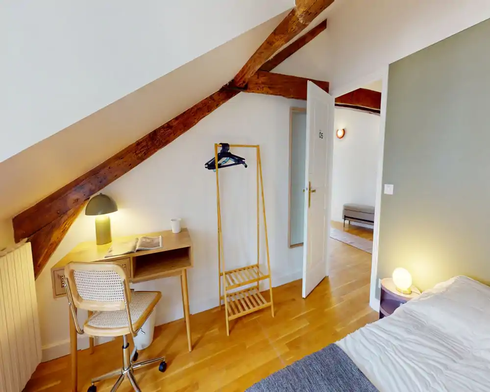 Bright, cozy attic bedroom with exposed wooden beams, a bedside table and lamp, a compact desk and an open clothes rack — good for a single occupant.
