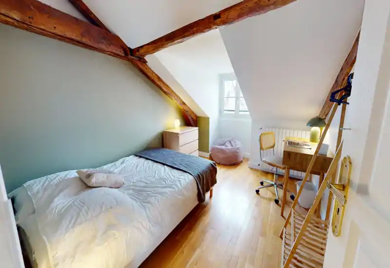 Sunlit attic bedroom with exposed wooden beams, a neatly made single bed, a small desk and a cozy beanbag — ideal for a quiet study or restful stay.