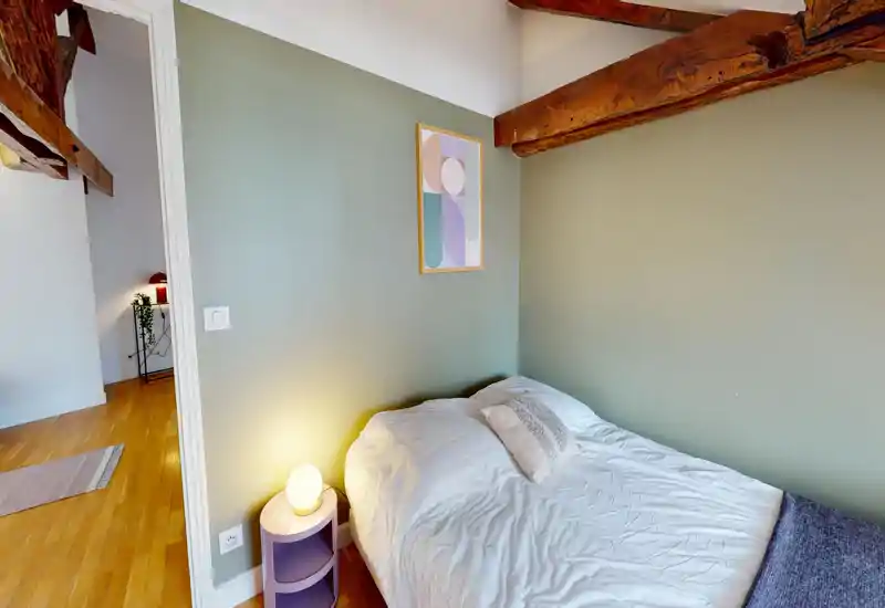 Cozy attic bedroom with exposed wooden beams, a neatly made double bed pushed against a soft green wall and a warm bedside lamp — ideal for a restful stay.
