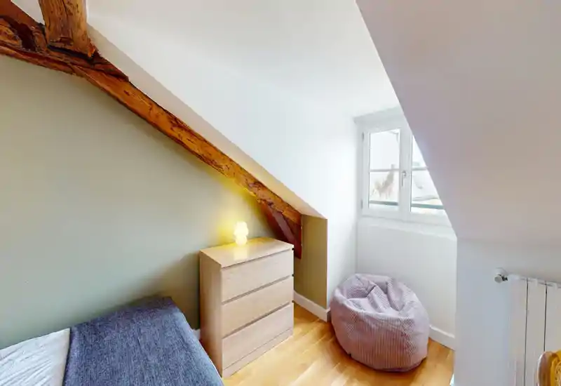 Small attic bedroom with exposed wooden beam, a single bed partially visible, a light wood chest of drawers and a cozy beanbag by a bright window.