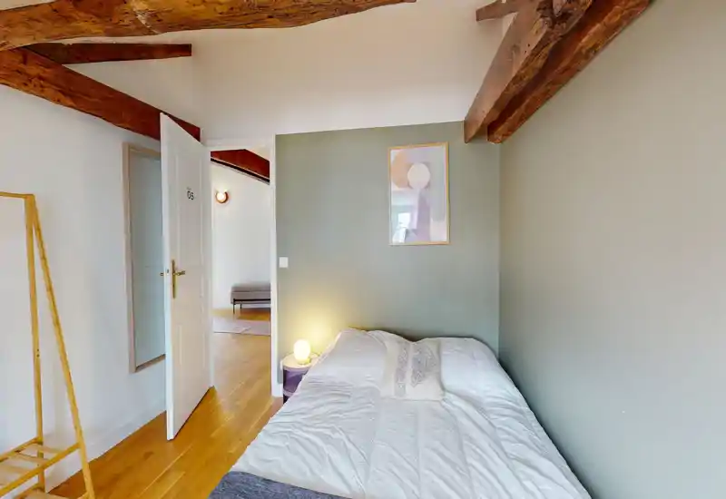Bright, cozy small bedroom with a comfortable double bed pushed against a green accent wall, warm wooden ceiling beams and a bedside lamp creating a welcoming atmosphere.