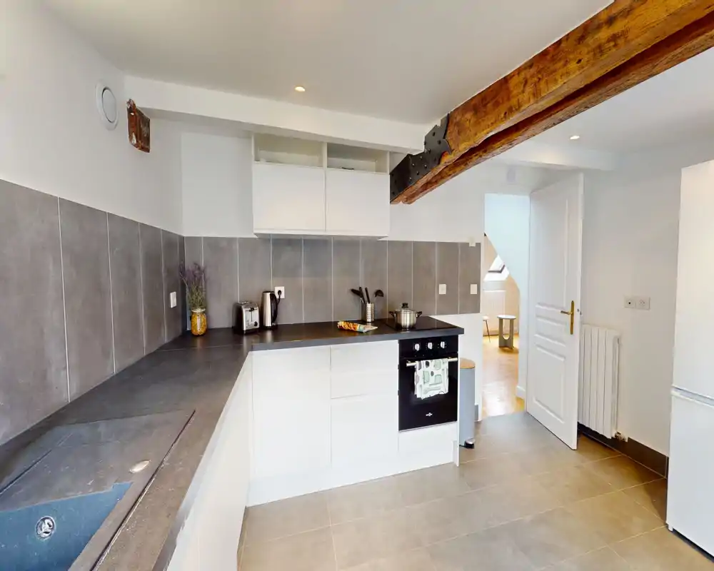 Modern, compact kitchen with white cabinetry, integrated oven, dark countertop and exposed wooden beam; clean and well lit, ideal for an apartment listing.